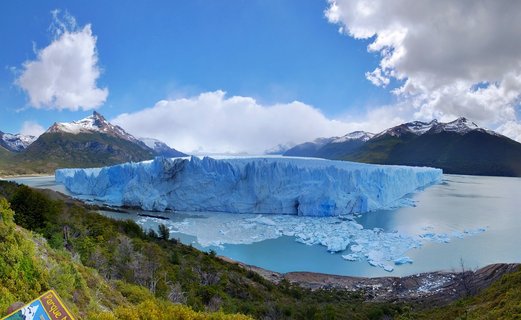 Massive blue glacier wall extending across turquoise lake water with snow-capped mountains and cloudy sky in Patagonia, Argentina