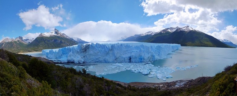 Massive blue glacier wall extending across a turquoise lake with floating ice chunks, snow-capped mountains and green forest in the foreground under a partly cloudy blue sky