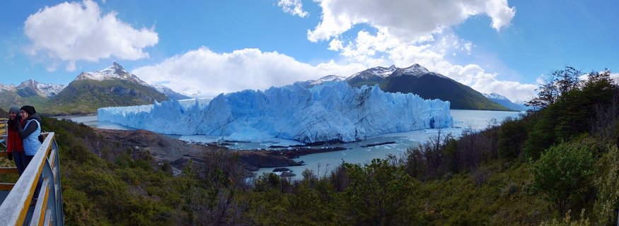 Panoramic view of the Perito Moreno Glacier's blue ice wall extending across Lago Argentino, with two tourists on a wooden viewing platform at left, green Patagonian vegetation in foreground, and snow-capped mountains under a partly cloudy sky
