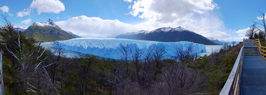 Panoramic view of Perito Moreno Glacier showing massive blue ice wall extending across Lago Argentino with mountains, forest, and observation walkway visible