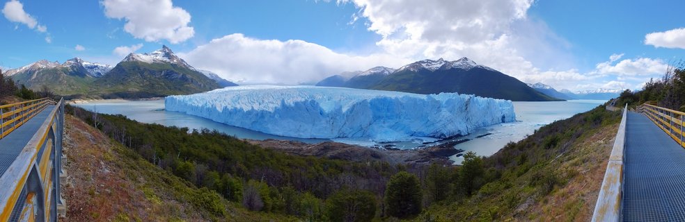 Panoramic view of the Perito Moreno Glacier showing massive blue ice formation in lake water with snow-capped mountains, green vegetation, and yellow viewing platforms on both sides under partly cloudy sky