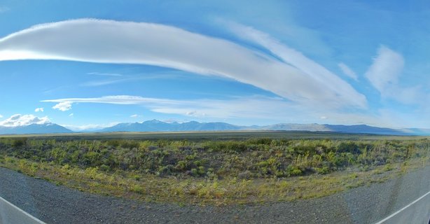 Wide landscape photograph showing Patagonian steppe vegetation in foreground, distant Andes mountains, and elongated white lenticular clouds against blue sky