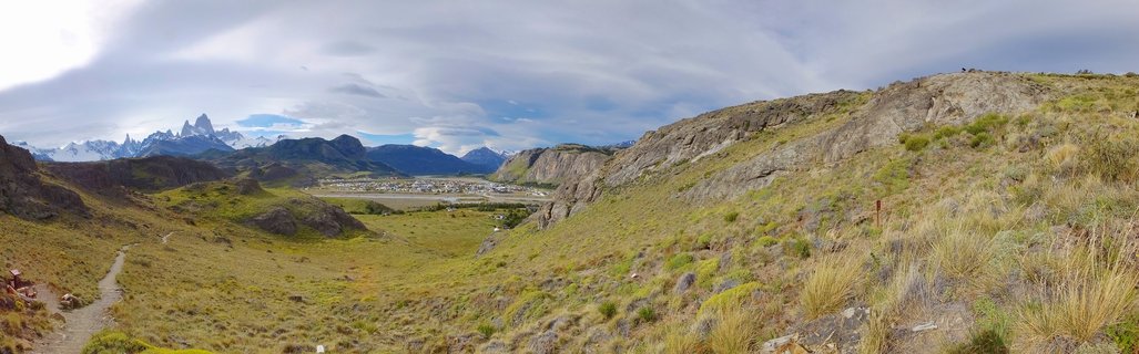 Panoramic landscape showing the town of El Chaltén in a valley with Mount Fitz Roy mountain peaks in the distance, rocky hillsides and grassy terrain in foreground, Patagonia Argentina