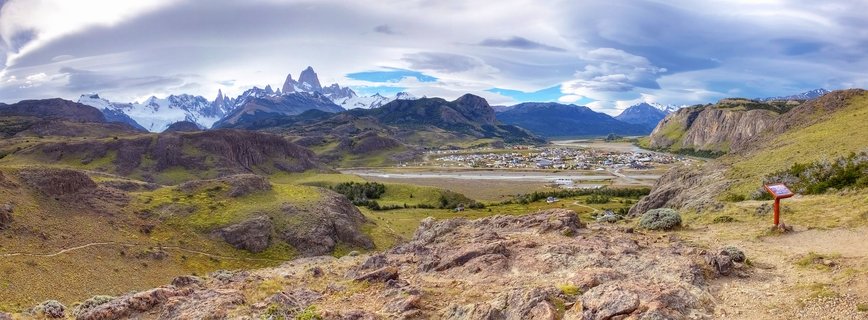 Panoramic landscape showing Mount Fitz Roy's jagged snow-capped peaks rising above the town of El Chaltén in a green valley, with rocky foreground terrain and dramatic cloudy sky in Patagonia, Argentina