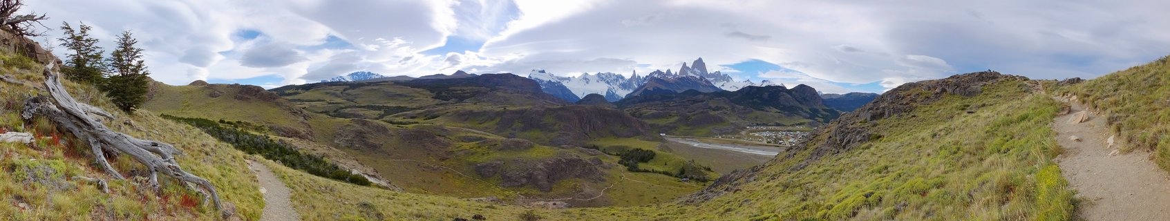 Panoramic landscape showing the jagged snow-capped peaks of Mount Fitz Roy mountain range in the distance, with green and brown grassy hillsides in the foreground, hiking trails on both sides, and a river valley below under a cloudy sky