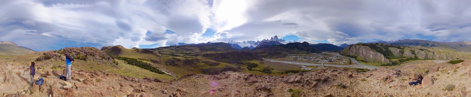 Panoramic landscape showing the Fitz Roy mountain range in the distance with dramatic clouds, a valley with a small town and winding river in the middle ground, and rocky terrain with several hikers in the foreground