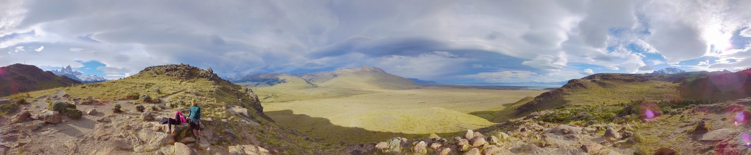 Two hikers sitting on rocky terrain overlooking a vast green valley with mountains in the distance under cloudy sky in Patagonia, Argentina