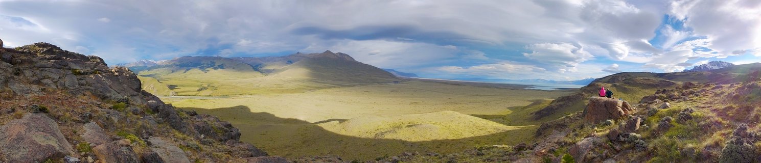 Panoramic landscape showing two hikers sitting on rocky outcrop overlooking wide Patagonian valley with mountains, grasslands, and dramatic cloud shadows near El Chalten, Argentina