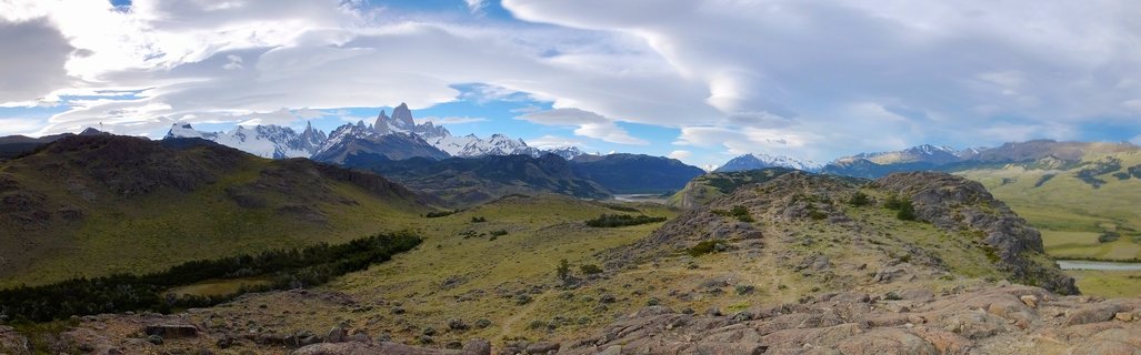 Panoramic view of Mount Fitz Roy's jagged snow-capped peaks rising above rolling green and brown Patagonian hills under a dramatic cloudy sky