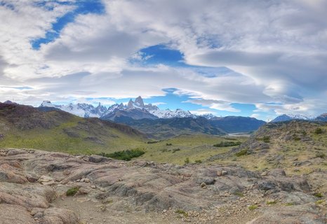 Panoramic landscape showing Mount Fitz Roy mountain range with snow-covered jagged peaks rising above green and brown Patagonian grasslands, rocky foreground terrain, and dramatic cloudy sky with blue patches