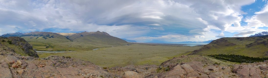 Wide landscape photograph showing rocky terrain in foreground, green valley with river in middle ground, and mountain range with dramatic cloudy sky in background near El Chalten, Argentina