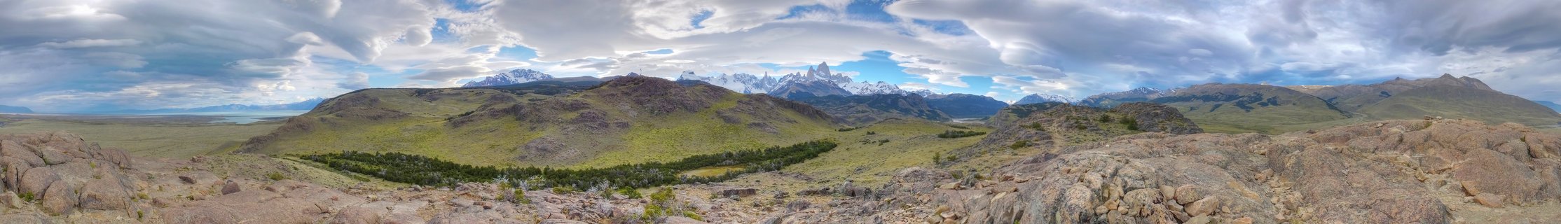 Wide panoramic landscape showing Fitz Roy mountain range with snow-capped peaks in the distance, green Patagonian steppe in the middle ground, and rocky terrain in the foreground under cloudy sky