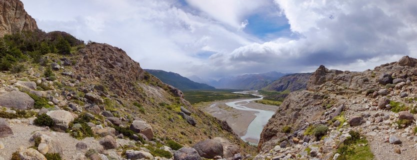 Wide panoramic landscape showing a rocky mountain valley with a winding glacial river, sparse vegetation, boulders in the foreground, and distant mountains under cloudy sky in El Chaltén, Argentina