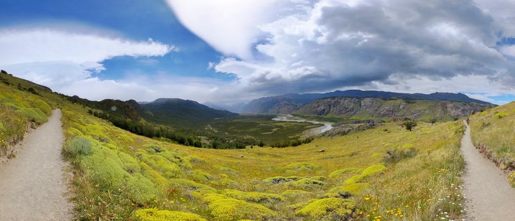 Wide landscape panorama showing a green valley with winding river, mountains in background, hiking trails on both sides, dramatic clouds overhead, El Chaltén, Argentina