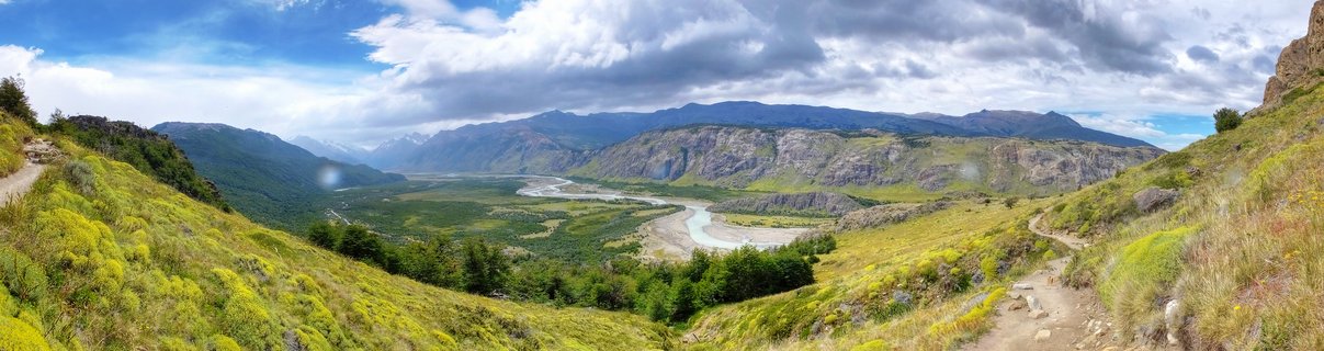 Panoramic mountain landscape showing a wide valley with a winding braided river, green and yellow vegetation on hillsides, distant mountain ranges under a cloudy sky, with hiking trails visible on both slopes