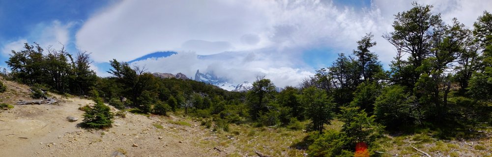 Mountain peak partially obscured by clouds rises above green forest landscape with dirt trail in Patagonia, Argentina