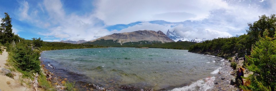 Panoramic landscape showing Mount Fitz Roy's sharp granite peak rising above turquoise Laguna de los Tres, with Patagonian forest along the shoreline and hikers visible on the right side under a partly cloudy sky