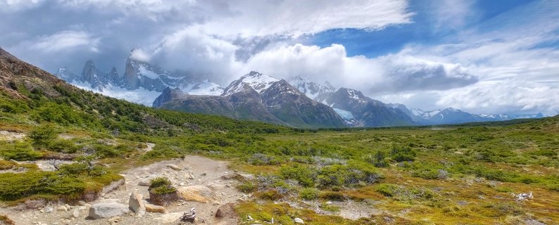 Wide landscape photograph showing a dirt hiking trail through green and yellow vegetation leading to snow-covered mountain peaks under a blue sky with white clouds