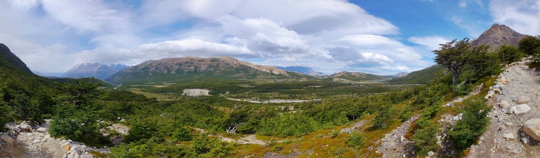 Panoramic landscape photograph showing mountain ranges and green valley in Patagonia near El Chalten, Argentina, with rocky foreground terrain, native lenga trees, blue sky and white clouds during summer afternoon