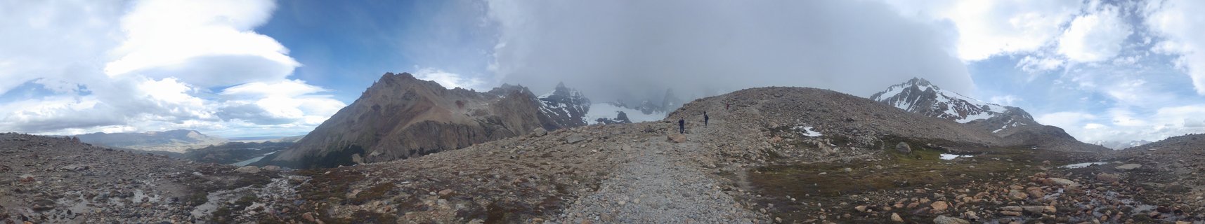 Panoramic landscape showing two hikers on a gravel path through rocky terrain with snow-capped mountains and cloudy sky in El Chalten, Argentina
