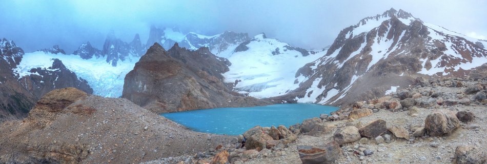 Panoramic landscape showing a bright turquoise alpine lake in front of snow-covered mountain peaks and glaciers under an overcast sky, with rocky terrain in the foreground