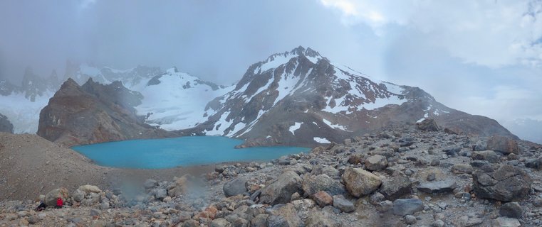 Turquoise glacial lake surrounded by rocky terrain with snow-capped mountain peaks and glaciers in the background under cloudy sky in El Chaltén, Argentina