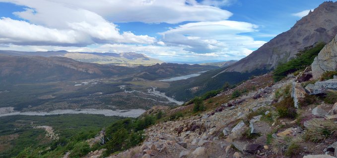 Mountain valley landscape showing a braided river flowing through green vegetation, with a lake and rugged peaks under a blue sky with white lenticular clouds