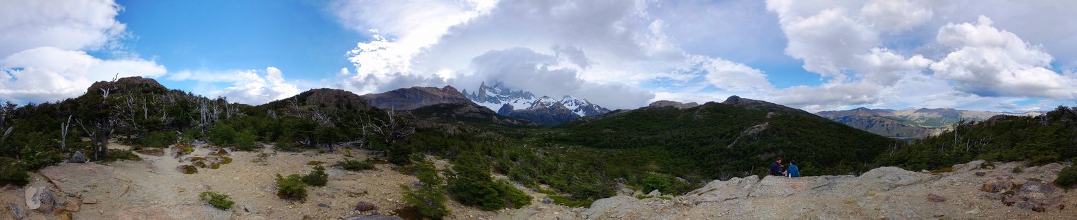 Panoramic view of snow-capped Mount Fitz Roy mountain peaks rising above green hills and rocky foreground terrain with two hikers visible, El Chaltén, Patagonia, Argentina