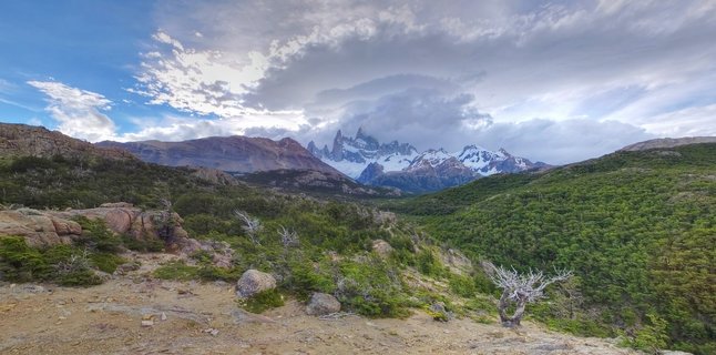 Panoramic landscape showing snow-capped Mount Fitz Roy mountain peaks rising above green forested hills with rocky foreground terrain and dramatic cloudy sky in El Chaltén, Argentina