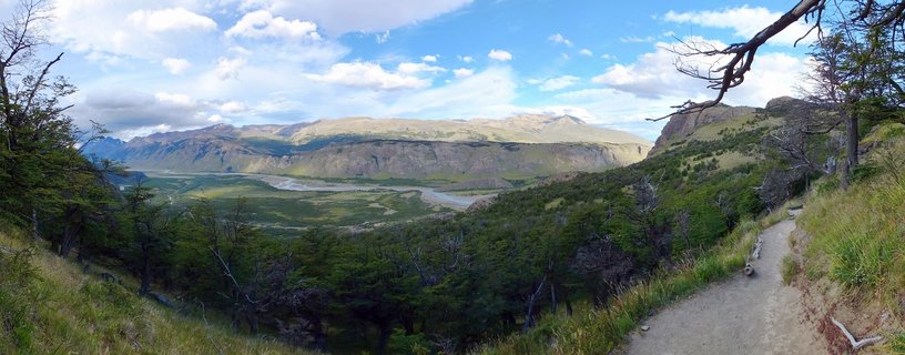 Panoramic landscape showing a green valley with winding river, forested hillsides, and mountains under blue sky with clouds in Patagonia, Argentina