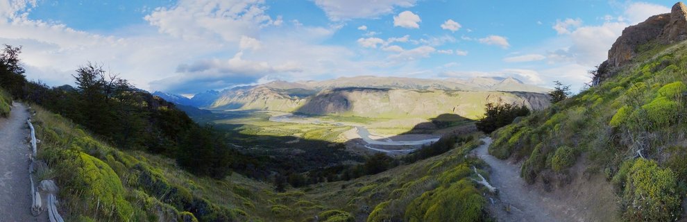 Wide landscape view of a green Patagonian valley with a winding river, mountain ranges in the distance, hiking trails on hillsides, and blue sky with white clouds above El Chalten, Argentina