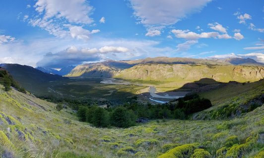 Wide landscape photograph of a Patagonian valley with a winding braided river, grassy hillsides with low vegetation, forest patches, and mountain ranges under a blue sky with scattered clouds