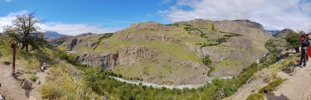 A panoramic view of the rugged Patagonian landscape near El Chaltén, Argentina, featuring a winding glacial river cutting through rocky hillsides covered in sparse vegetation under a blue sky with scattered clouds.