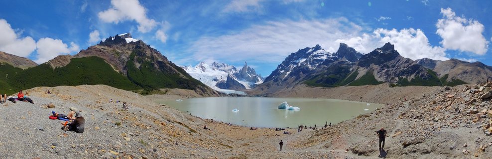 Panoramic view of hikers on rocky terrain beside a glacial lake with Mount Fitz Roy and snow-capped peaks in the background under a blue sky with scattered clouds in El Chaltén, Argentina