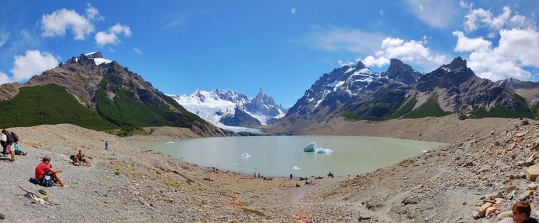 Wide mountain landscape showing hikers sitting on rocky terrain beside a glacial lake with icebergs, snow-capped Fitz Roy peaks and glaciers in the background under blue sky with clouds
