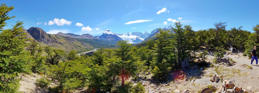 Panoramic view of snow-capped Fitz Roy mountain range in Patagonia with green forest foreground and hikers on a trail under blue sky