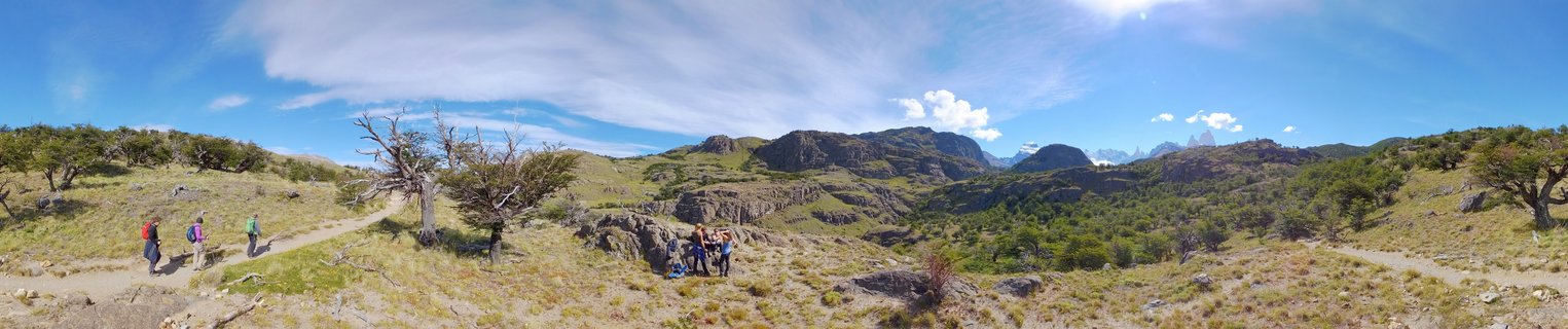 Group of hikers with colorful backpacks walking on a dirt trail through grassy Patagonian steppe with windswept trees, rocky hills, and distant mountain peaks under blue sky