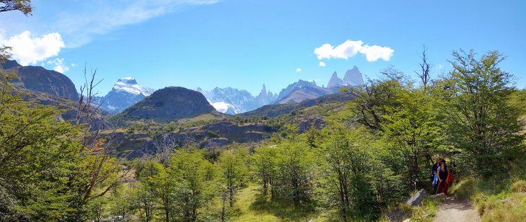 Panoramic view of snow-capped Mount Fitz Roy mountain range in Patagonia with green forest in foreground and two hikers on a trail