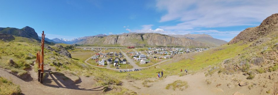 Panoramic view of El Chaltén village in a green valley surrounded by rocky mountains and snow-capped peaks under blue sky with hikers on foreground trail