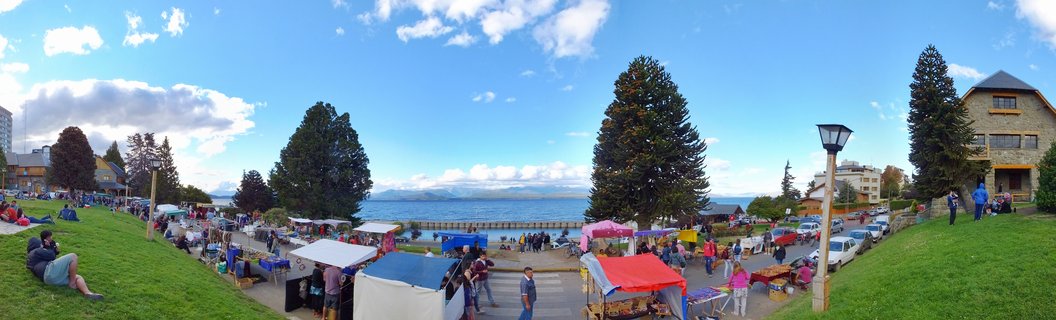 Outdoor artisan market with colorful tents and people along a grassy lakeshore, with blue water and mountains in the background under a partly cloudy sky in Bariloche, Argentina