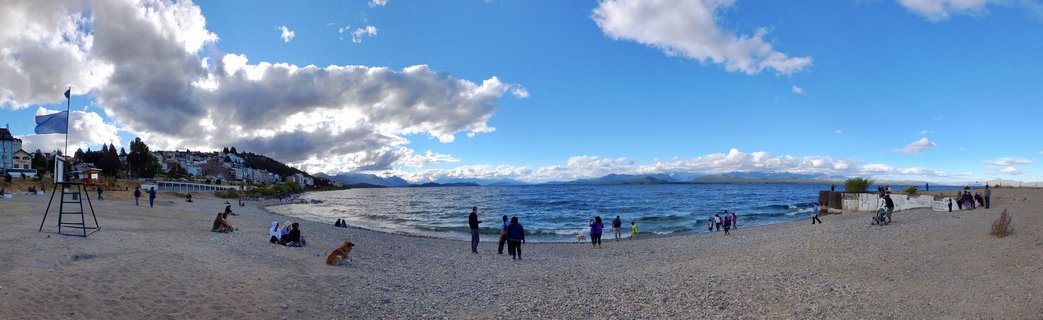 Wide panoramic view of a pebble beach along a large lake with people scattered across the shore, mountains visible in the distance under a blue sky with white clouds