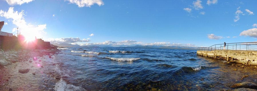 Wide lakeside panorama showing blue water with small waves, a concrete pier with metal railings on the right where a person stands, rocky beach on the left, distant mountains across the water, and bright sun with lens flare in a blue sky with scattered clouds