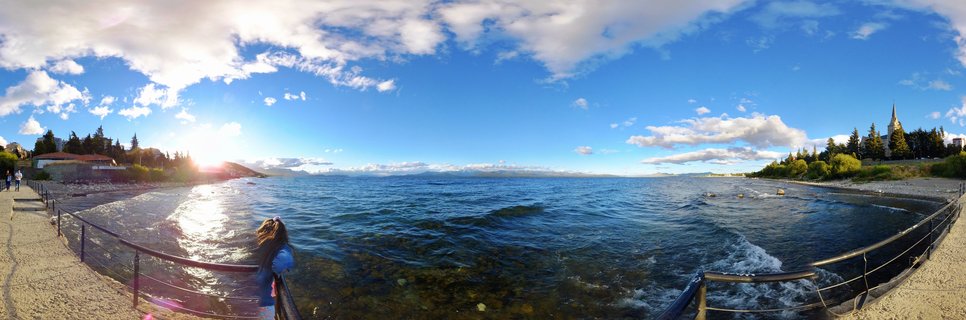 Panoramic lakeside scene showing a person at a railing overlooking blue water with sun reflection, stone cathedral on right shoreline, trees, distant mountains, and cloudy sky at golden hour in Bariloche, Argentina