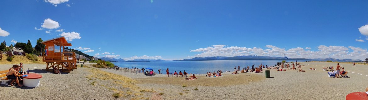 Wide beach scene showing people sunbathing and relaxing on pebble shore of Lake Nahuel Huapi with wooden lifeguard tower and distant mountains under blue sky