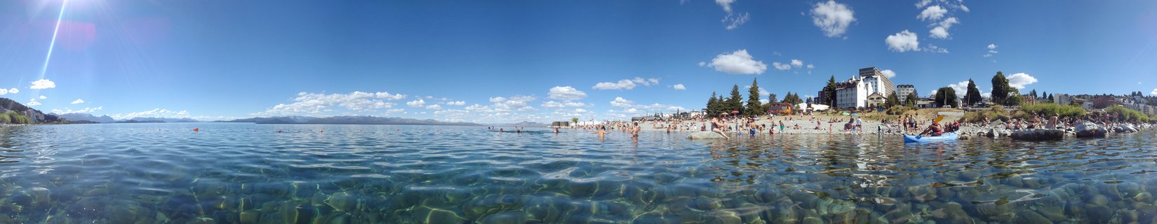 Panoramic view of a crowded beach at a clear blue lake with mountains in the distance, people swimming in transparent water, buildings along the shoreline under a bright blue sky with scattered clouds