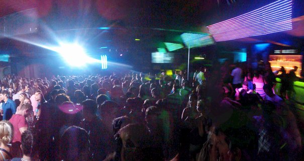 Large crowd of people at indoor nightclub with bright stage spotlight and colorful LED ceiling light panels in purple, blue and green