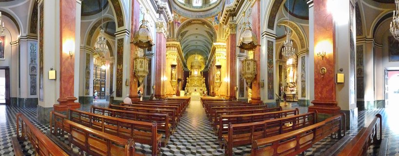 Interior view of a cathedral with wooden pews, red marble columns, golden altarpiece, chandeliers, and checkered black and white floor