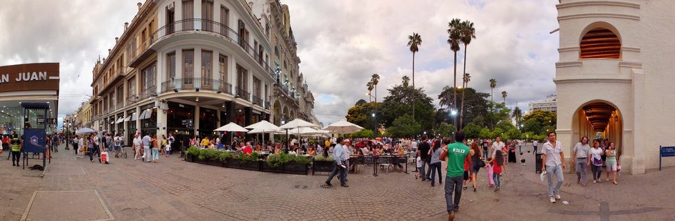 Wide panoramic street view of a colonial plaza in Salta, Argentina showing historic buildings with balconies, a white church tower with arches, palm trees, outdoor café seating with white umbrellas, and numerous pedestrians on cobblestone pavement under cloudy sky