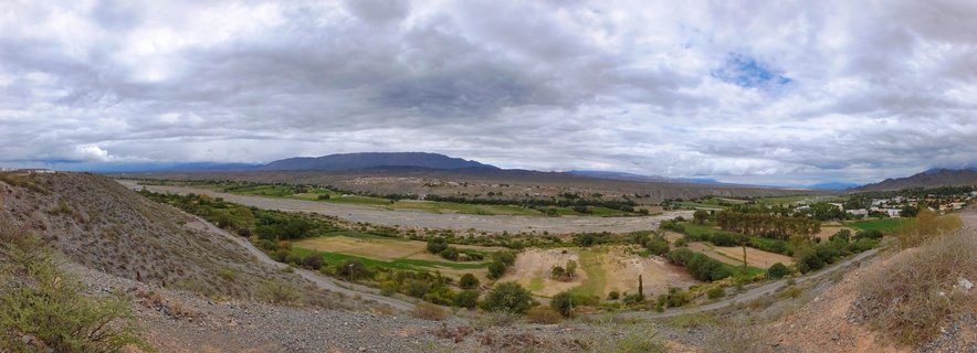 Wide panoramic landscape of a valley in Salta, Argentina showing green agricultural fields, a winding river, distant blue mountains under an overcast gray sky with layered clouds, and sparse vegetation on arid foreground hillsides