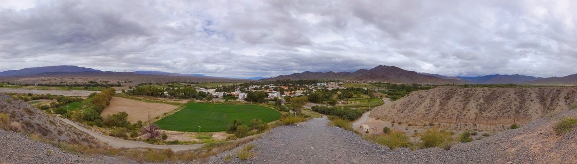 Panoramic landscape photograph of Salta, Argentina showing a wide valley with green agricultural fields including a circular irrigated field, a small town with white buildings in the middle distance, arid rocky hills and mountains in the background, and dramatic overcast sky cover